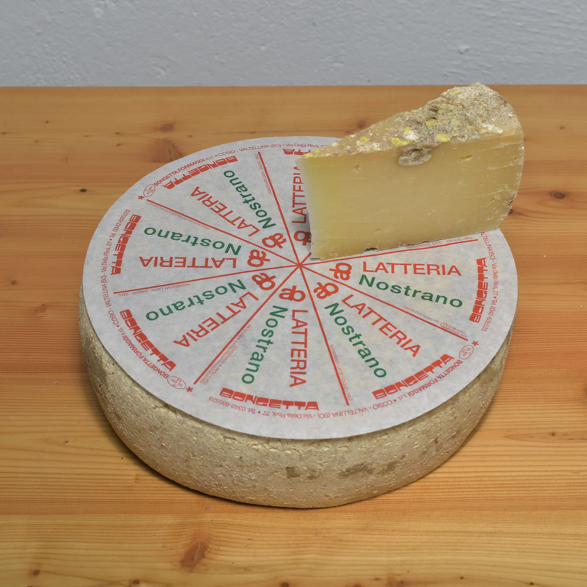 Wheel of Latteria Nostrano on Bongetta Formaggi’s printed paper, Cosio Valtellino; a cut wedge shows the firm paste with fine eyes.